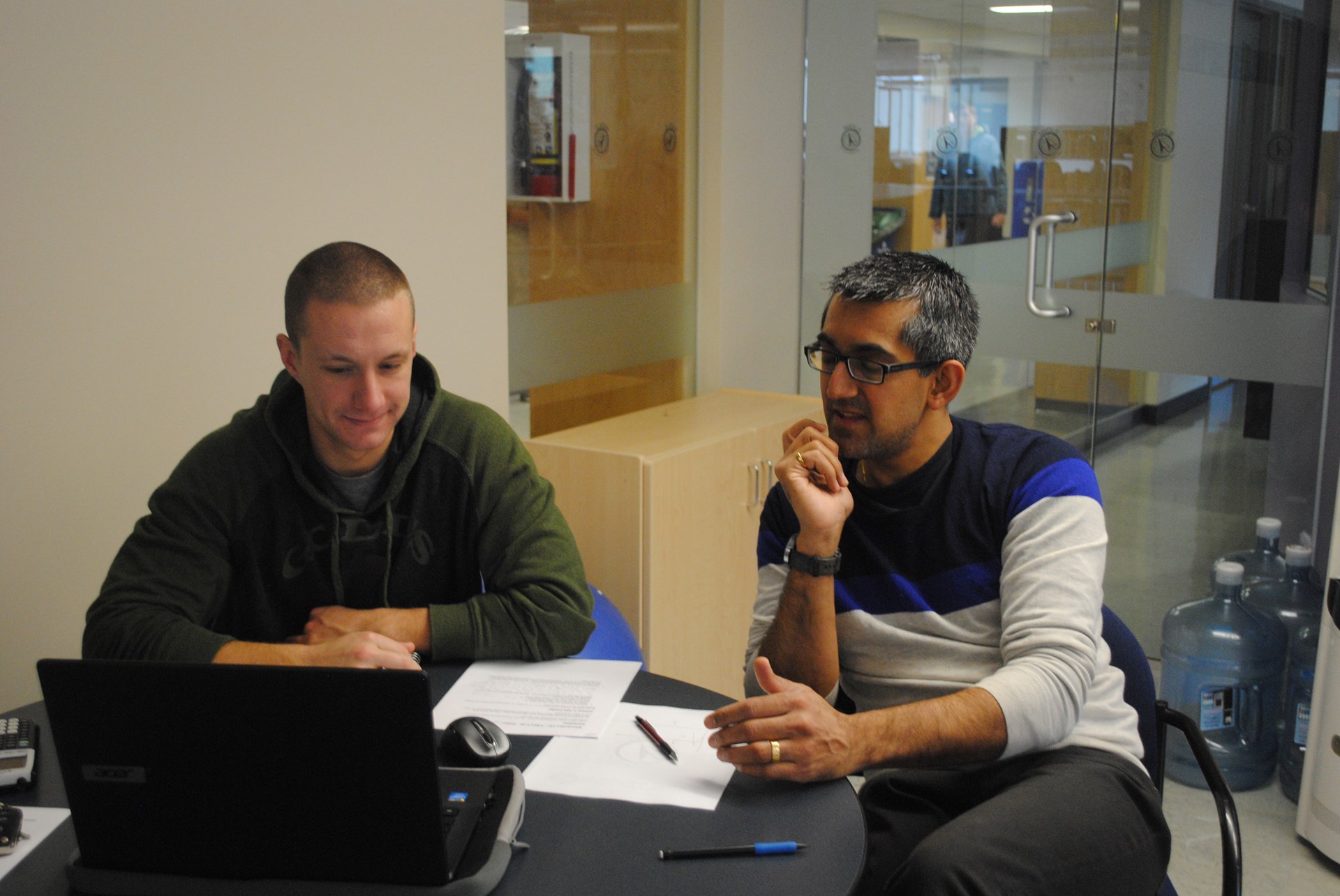 A student and teacher sitting in a school work-space, looking at notes and a laptop together, teacher explaining something.