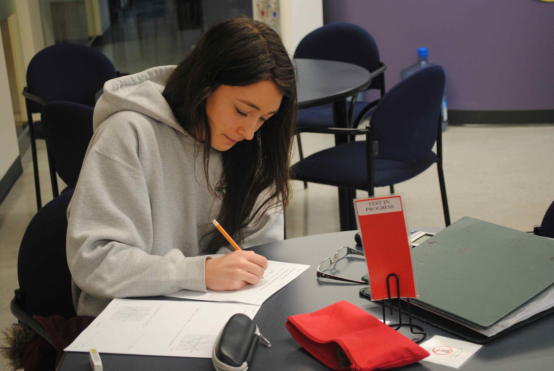 A student sitting alone at a round table in a school setting, making notes with a pencil