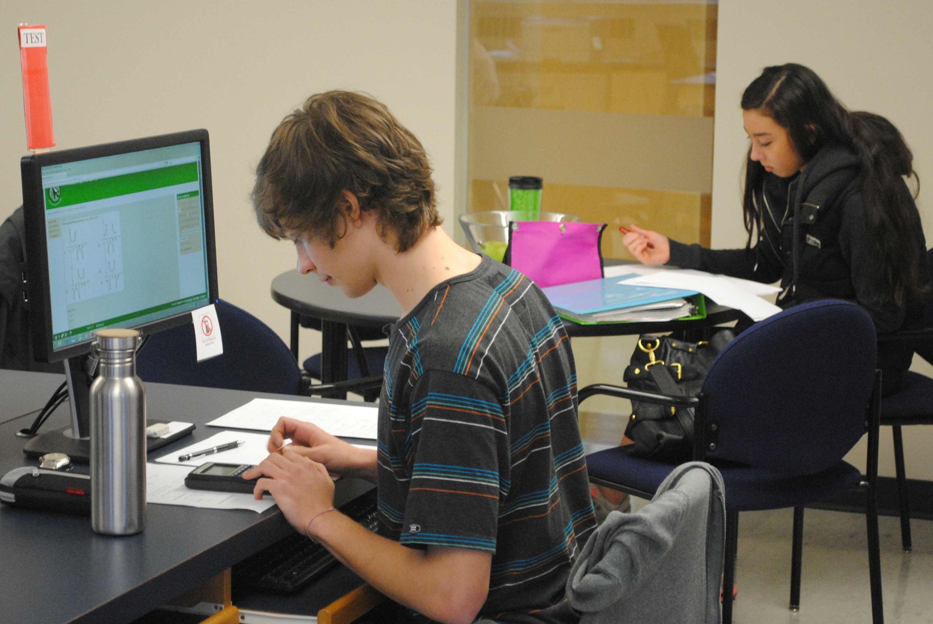 a student working at NVOL on a computer, another in the background working at a table, on paper