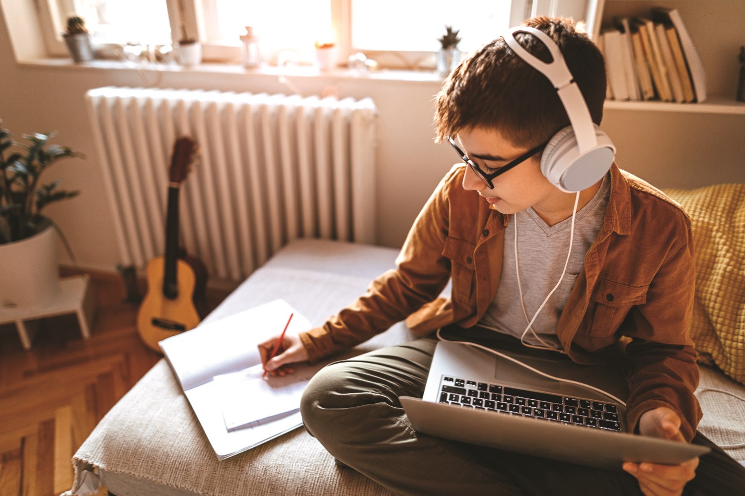 A student at home on a comfortable chair, wearing headphones, with a laptop on lap and notebook beside, making notes.